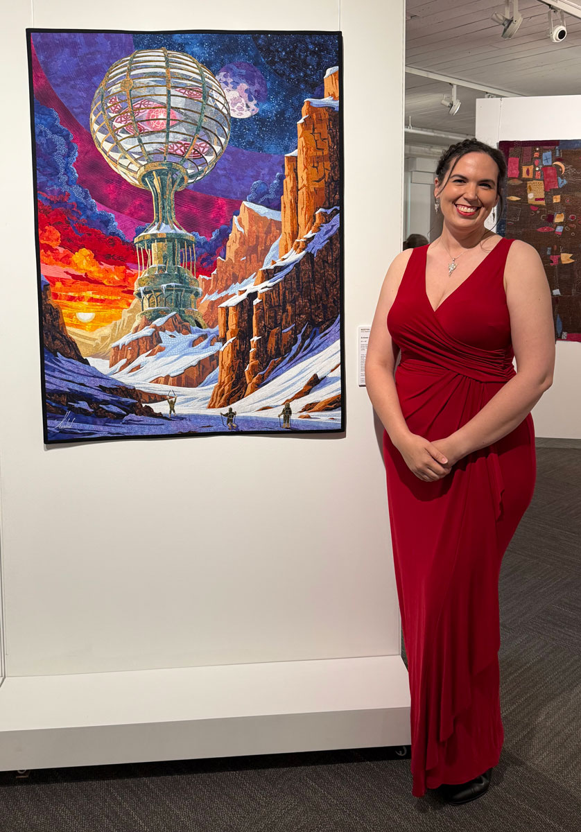 Kestrel dressed in a red evening gown and standing next to her quilt